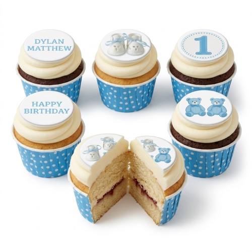 Assortment of baby boy birthday cupcakes with blue polka dot wrappers, white frosting, and edible toppers featuring baby booties, teddy bears, and a number one.