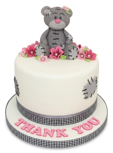 White fondant cake with grey teddy bear topper, pink flowers, and stitched patches. Custom message board.