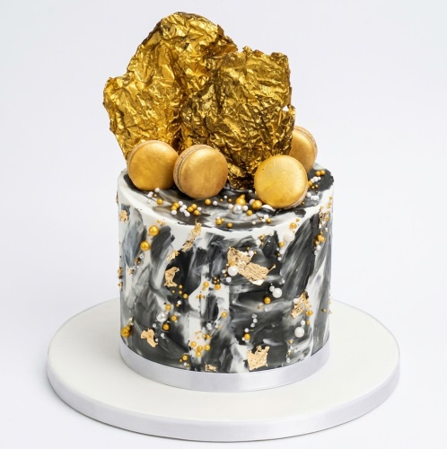 Single-tier black and white marble cake with gold foil, gold macarons, and sprinkles.
