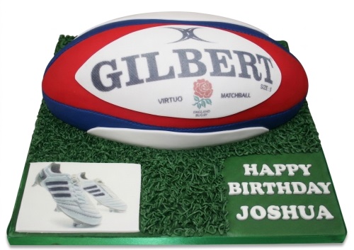 3D rugby ball cake on a green grass-effect board with edible rugby boots and a personalised message.