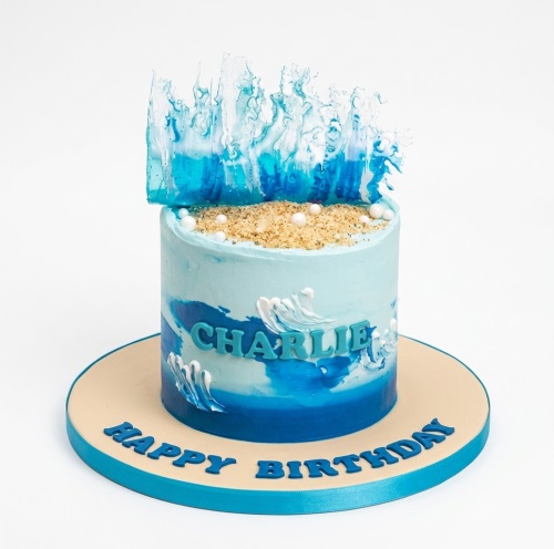 Single-tier blue ocean theme birthday cake with edible waves, sand, and white pearls, on a beige board.