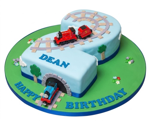 Light blue number two cake with red and blue toy trains on tracks, green base, and personalised message.