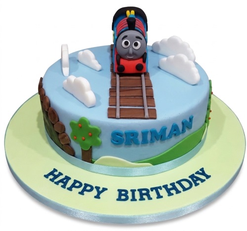 Round blue train theme birthday cake with 3D locomotive, tracks, clouds, and green hills.
