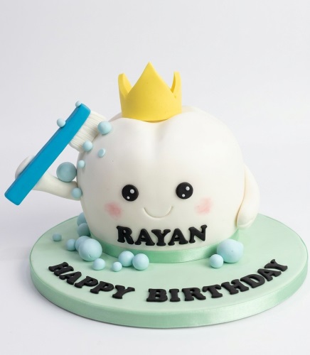 White tooth-shaped cake with a yellow crown, blue toothbrush, and bubbles on a green board.