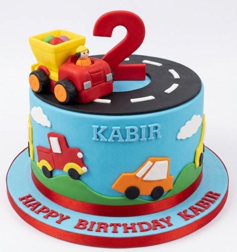 Blue birthday cake with red and orange cars, a truck, road, and a red custom age topper for a boy's birthday.