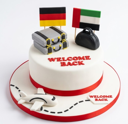 White welcome back cake with red ribbon, German and UAE flags, grey suitcases, black handbag, and a white airplane.