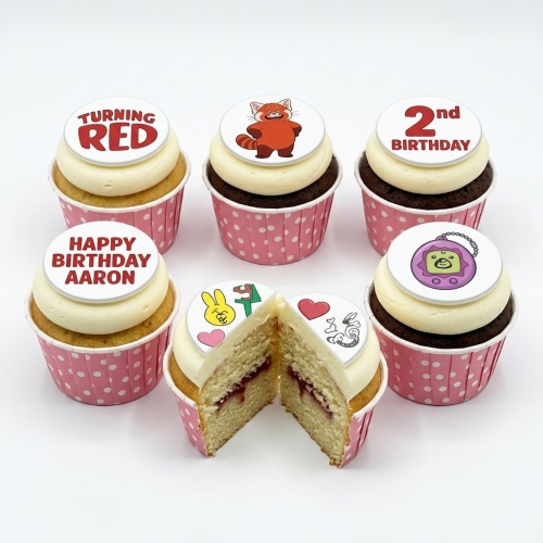 A set of six Turning Red themed birthday cupcakes with white frosting and pink polka dot cases, featuring edible toppers including a red panda, 'Turning Red' logo, '2nd Birthday', 'Happy Birthday Aaron', and a Tamagotchi-like toy. One cupcake is cut open revealing a jam filling.
