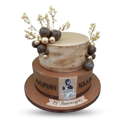 Two-tier brown and beige anniversary cake with Alton Ellis image, "15th Anniversary" banner, gold sprigs, and chocolate-gold spheres.