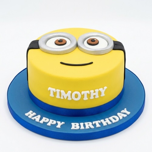 Round yellow Minion character birthday cake with goggles and a smile, on a blue cake board.