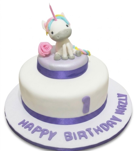 White fondant unicorn birthday cake with a rainbow mane, pink rose, and purple ribbon, featuring a custom age number.