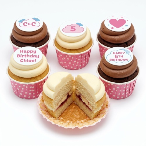 Assortment of vanilla and chocolate cupcakes with white and brown frosting, pink polka dot cases, and customisable birthday toppers including age, initials, and heart designs.