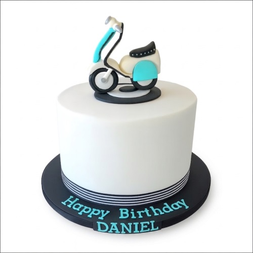 White round birthday cake with a light blue and white scooter topper and a personalised message board.