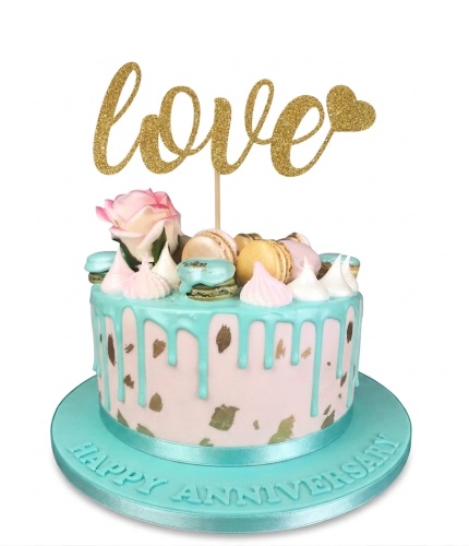 Round anniversary cake with mint green drip, gold speckles, macarons, meringue, pink rose, and a gold 'love' topper.