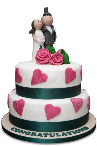 Two-tier white wedding cake with bride and groom toppers, pink roses, green ribbon, and pink hearts.