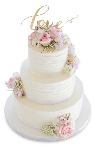 Three-tiered white wedding cake with pink roses, white flowers, and a gold 'love' topper.