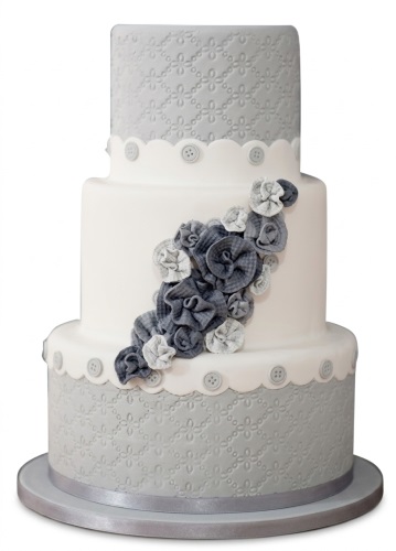 Three-tiered white and grey cake with fabric-style floral decorations and button details.