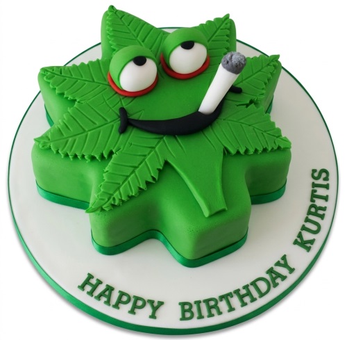 Green cannabis leaf shaped birthday cake with cartoon eyes, a smile, and a lit joint, on a white board with a custom message.