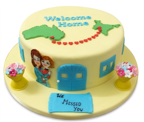 Round yellow Welcome Home cake with maps of Australia and India, red heart trail, woman and child figures, blue door, and flower pots.