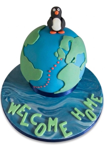 3D globe cake with green landmasses, a small black and white penguin topper, and a blue marbled cake board with light green lettering.