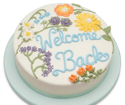 Round white cake with 'Welcome Back' in blue script, decorated with orange, yellow, white, and purple piped flowers and green vines.