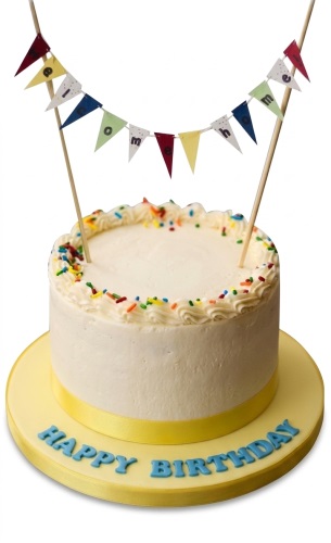 Round white birthday cake with colourful sprinkles, yellow ribbon, blue 'Happy Birthday' text, and a multicoloured 'Welcome' bunting banner.