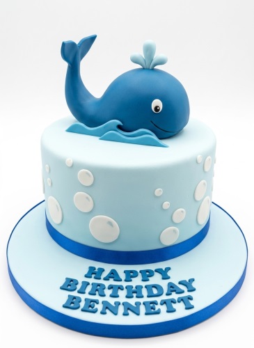 Light blue single-tier cake with a blue whale topper, waves, and white bubbles, with a personalised birthday message on the board.