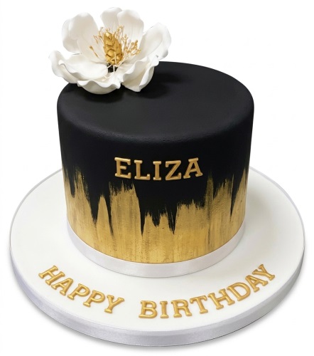 Round black cake with gold brushstrokes, white sugar flower, and 'Happy Birthday' message.