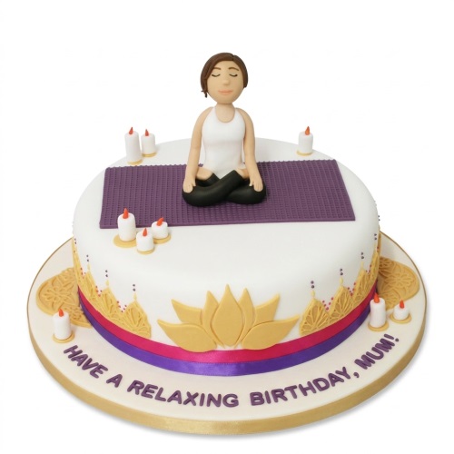 Round white cake with a yoga figure on a purple mat, candles, gold lotus, and ribbons.