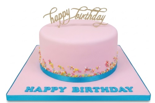 Round pink birthday cake with multicoloured sprinkles, blue ribbon, and a gold 'Happy Birthday' topper.